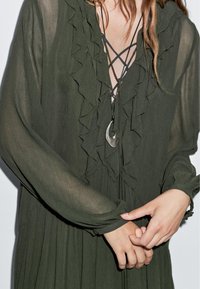 Olivengrünes Kleid mit Rüschendetails am Ausschnitt und den Ärmeln. Transparenter Stoff und ein plissierter Rock, ergänzt durch eine Anhängerhalskette.