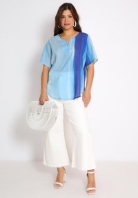 Blauwe gradient blouse met korte mouwen en een keyhole halslijn, gecombineerd met witte wijde broek en een witte gevlochten tas. De hakken zijn wit met enkelbandjes.