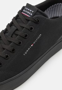 Baskets noires en toile avec bout rond, logo "TOMMY HILFIGER" blanc et accent rouge/blanc/bleu sur la languette, dotées de lacets et d'œillets noirs.