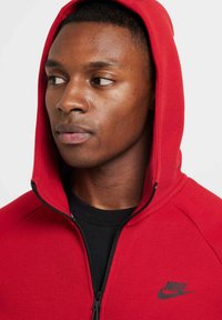 Sudadera roja con cremallera hecha de una mezcla de algodón, con el logotipo negro de Nike en el pecho. Presenta una capucha forrada y un cuello acanalado.