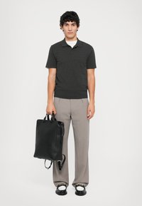 Zaino in pelle nera con manici doppi e spallacci; modello in polo grigia scura e pantaloni grigi chiari, in piedi su uno sfondo bianco.