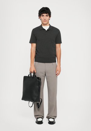 Zaino in pelle nera con manici doppi e spallacci; modello in polo grigia scura e pantaloni grigi chiari, in piedi su uno sfondo bianco.