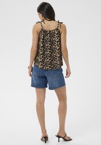 Mujer con top sin mangas de estampado de leopardo con tirantes de lazo, pantalones cortos de mezclilla y sandalias negras con tacón, de pie de espaldas sobre un fondo liso.