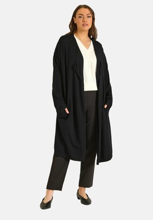 Frau trägt einen langen schwarzen offenen Cardigan, ein weißes V-Ausschnitt-Oberteil, schwarze Hosen und schwarze Schuhe, steht mit den Händen in den Taschen des Cardigans.