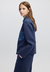 Marineblaues Sweatshirt mit einem Denim-Einsatz, das ein geometrisches Muster aufweist, entspannte Passform und seitliche Taschen hat, kombiniert mit passenden Hosen.