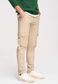 Homme portant un pantalon cargo beige, des baskets blanches, un sweat-shirt vert et une montre-bracelet, les mains dans les poches, sur un fond uni.