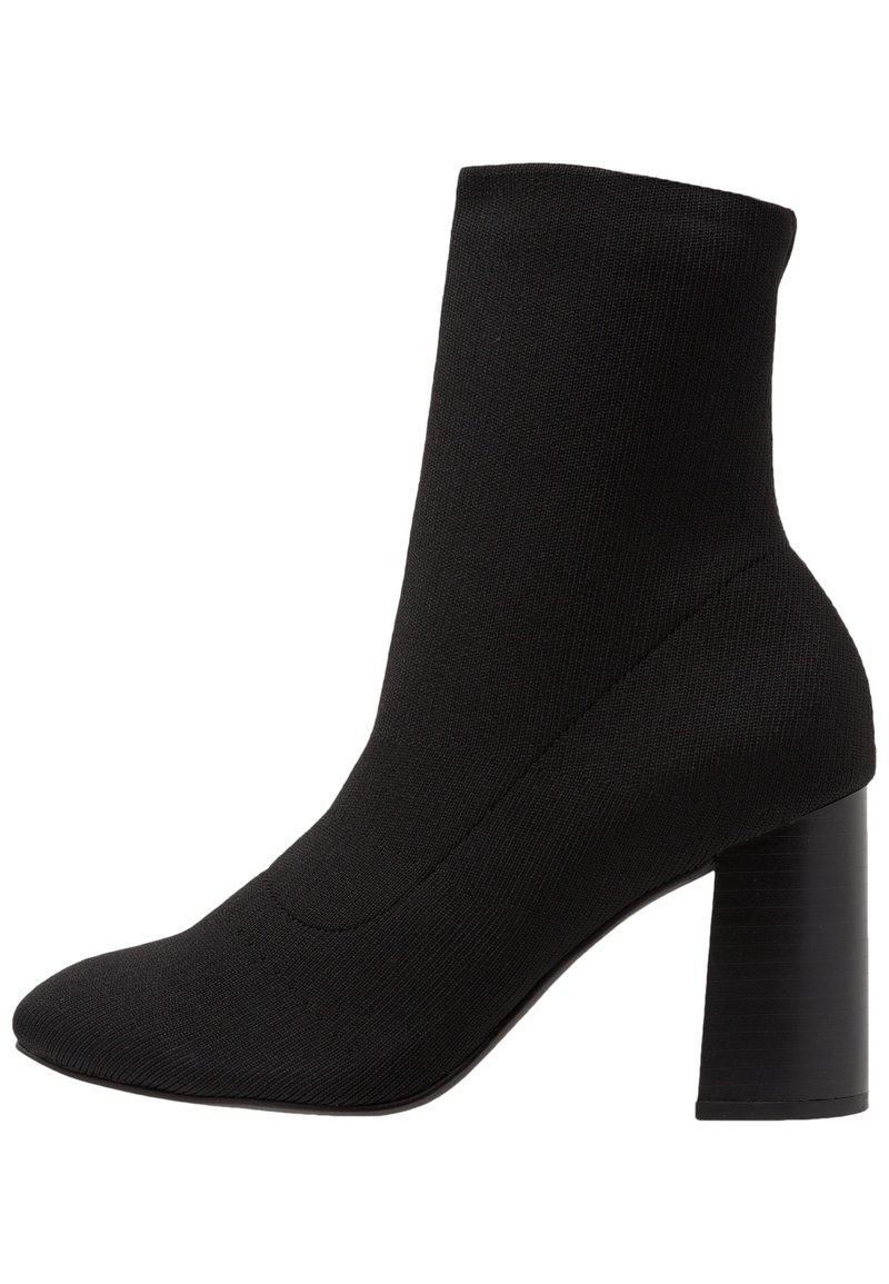 Bottine noire avec une tige en tricot ajustée, un bout pointu et un talon cylindrique épais. Texture lisse avec peu de détails.