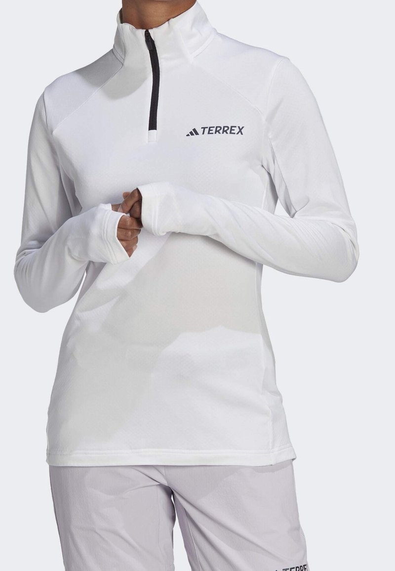 Pull-over de sport blanc à manches longues avec ouvertures pour les pouces et fermeture éclair noire, avec logo noir "TERREX" sur la poitrine.