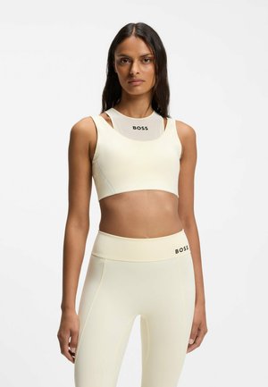Kvinde iført cremefarvet sportcroptop og leggings med "BOSS"-logo, stående foran en ensfarvet hvid baggrund.