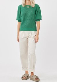 Personne portant un pull en maille à manches courtes vert, un pantalon plissé écru, des colliers en chaîne dorés et des sandales épaisses beige à enfiler.