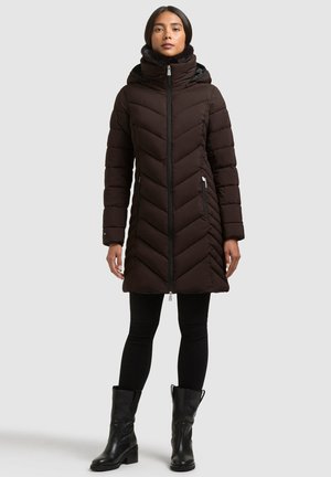 Une femme porte un manteau matelassé brun foncé, long jusqu’aux genoux et à col montant, un pantalon noir et des bottes noires, sur un fond uni.
