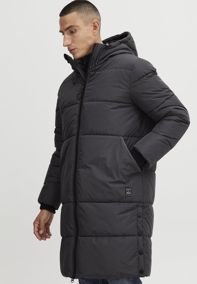 Manteau long noir rembourré avec capuche, présentant une fermeture éclair à l'avant, des poches latérales et des poignets élastiques. Détails de couture remarquables et patch logo.