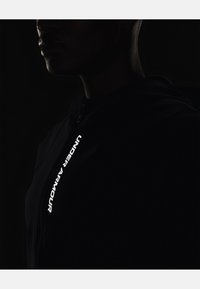 Čierna zipová mikina s vysokým golierom, na boku s výrazným bielym textom "UNDER ARMOUR". Hladká, elegantná tkanina s minimálnymi detailmi.