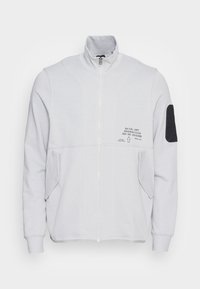 Ljust grå zip-jacka med hög krage, två fickor med lock, svart armbågsplåster på vänster ärm och liten svart text på vänster bröst.