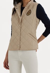 Béžový prošívaný vest s vysokým límcem, zdobený stříbrnými cvoky a vyšívanou nášivkou na prsou. Texturovaná tkanina, boční kapsy.