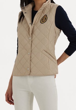 Béžový prošívaný vest s vysokým límcem, zdobený stříbrnými cvoky a vyšívanou nášivkou na prsou. Texturovaná tkanina, boční kapsy.