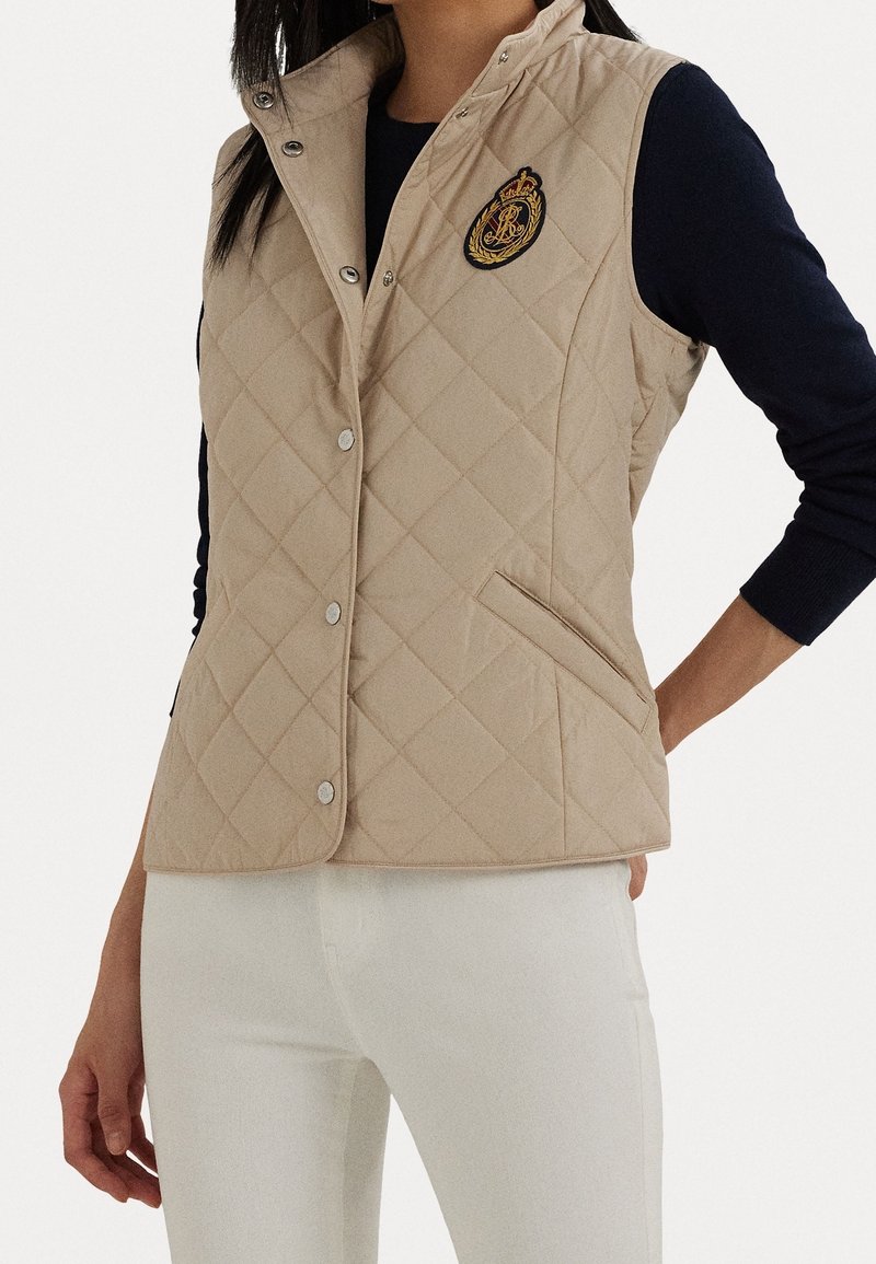 Béžový prošívaný vest s vysokým límcem, zdobený stříbrnými cvoky a vyšívanou nášivkou na prsou. Texturovaná tkanina, boční kapsy.