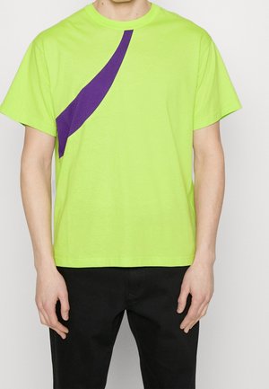 Mann trägt ein kurzärmliges, limettengrünes T-Shirt mit einem diagonalen lila Streifen, kombiniert mit schwarzen Hosen, und steht mit den Händen an den Seiten.