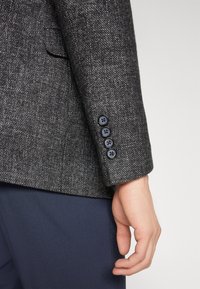 Manga de un blazer gris texturizado con cuatro botones negros y una textura de tela suave. Los pantalones azul marino son parcialmente visibles.