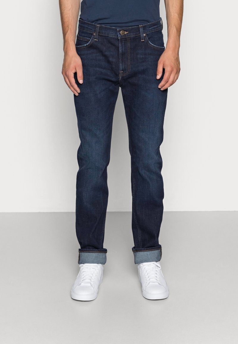 Mørkeblå denimjeans med rette ben, med oppbrettede kanter, fem lommer og en subtil kontrast søm detalj. Kombinert med hvite joggesko.