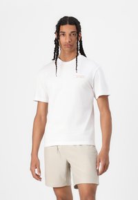 White cotton t-shirt with short sleeves, featuring a small pink logo on the left chest; paired with beige ribbed shorts.