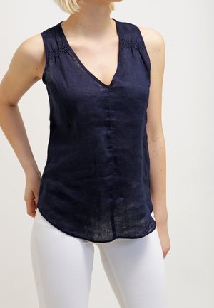 Kobieta ubrana w granatową bluzkę bez rękawów z dekoltem w serek i białe spodnie, stojąca z jedną ręką opartą na biodrze.