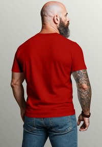 Red cotton t-shirt with short sleeves, fitted design. Visible tattoo on the left arm, worn with blue denim jeans. Plain background.