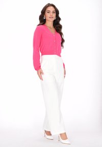 Élénk rózsaszín kardigán V-nyakkivágással és gombos részlettel, fehér, magas derekú, széles szárú nadrággal és fehér hegyes orrú cipővel párosítva.