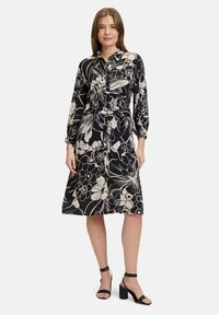 Svart och krämfärgad blommig klänning med långa ärmar, knäppning fram, bälte i midjan och knälång kjol. Texturerad tyg med djärva mönster.