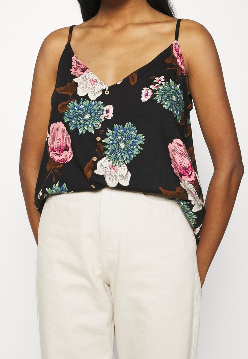 Femme portant un haut noir sans manches à motifs floraux roses, bleus et blancs, associé à un pantalon taille haute blanc cassé.