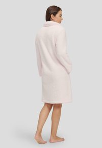 Bata de baño rosa claro, esponjosa, con cuello, mangas largas y bolsillos laterales, que presenta una textura suave y un diseño hasta las rodillas.