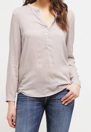Donna con camicetta rosa chiaro a maniche lunghe con piccolo motivo a pois e jeans blu, con una mano in tasca e l'altra al fianco.