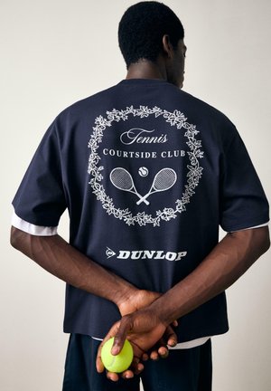 Homme en polo de tennis bleu marine avec le logo "Tennis Courtside Club" et raquettes croisées tient une balle de tennis derrière le dos.