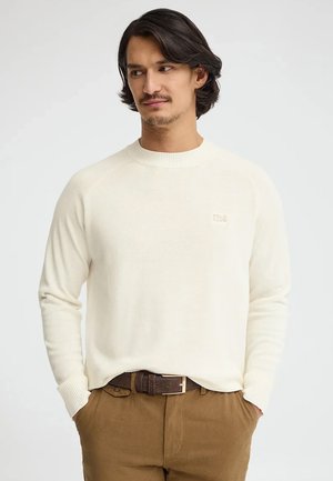 Uomo con capelli scuri che indossa un maglione crema a maniche lunghe e pantaloni marroni con cintura scura, mani nelle tasche, guarda di lato.