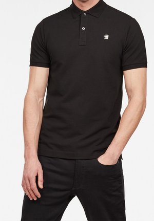 Mann trägt ein schwarzes Kurzarm-Poloshirt mit einem kleinen weißen Logo auf der Brust und schwarze Hose, eine Hand in der Tasche.
