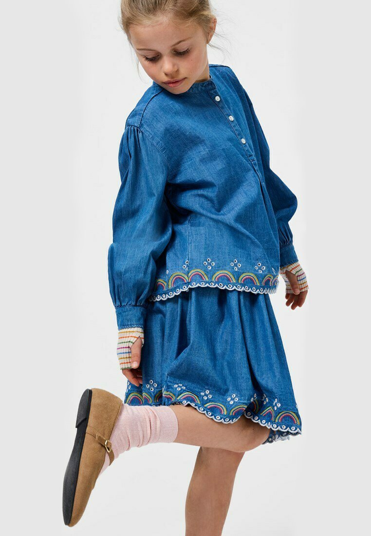 Ensemble robe en denim et blouse, avec des manches ballons, broderie arc-en-ciel, ourlet festonné et accents pastel. Chaussures brunes avec une chaussette rose.