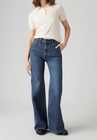 Krämfärgad kortärmad pikétröja i kombination med högmidjade mörkblå utsvängda jeans. Jeansen har en klassisk femficksdesign och lätt slitningar.