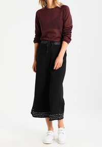 Sweat-shirt marron à manches longues, associé à un pantalon culotte noir avec un ourlet à pois. Chaussures blanches à texture.