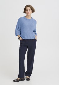 Pull bleu clair en tricot avec un col rond, associé à un pantalon large bleu marine et des chaussures foncées, créant une tenue décontractée et confortable.