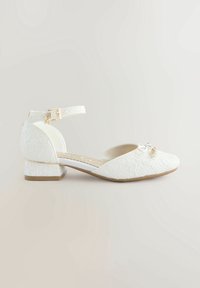 Chaussure blanche en dentelle à talon bas avec bride à boucle à la cheville et petit nœud décoratif sur le bout, sur un fond clair uni.