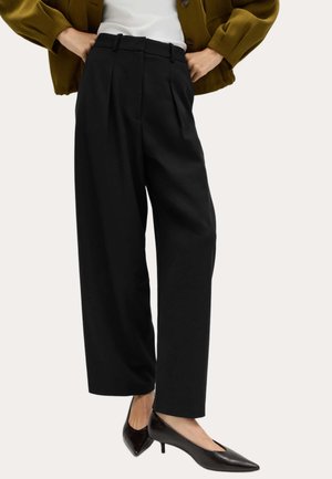 Vrouw draagt zwarte cropped wijde broek gecombineerd met zwarte puntige lage hak schoenen, witte top en een olijfgroene jas.