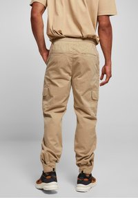Beige Cargohosen mit elastischem Bund, zulaufenden Bündchen und Seitentaschen. Getragen mit schwarzen und orangefarbenen Turnschuhen vor neutralem Hintergrund.