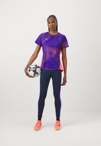 Violeta un rozā acs sporta krekls ar īsām piedurknēm, apvienots ar tumši ziliem legingiem. Modelis tur futbolu un valkā košas sporta kurpes.