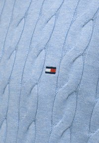 Ανοιχτό μπλε πλεκτό πουλόβερ με σχέδιο καλώδιο, με ένα μικρό λογότυπο σε κόκκινο, λευκό και μπλε στην κάτω αριστερή πλευρά.