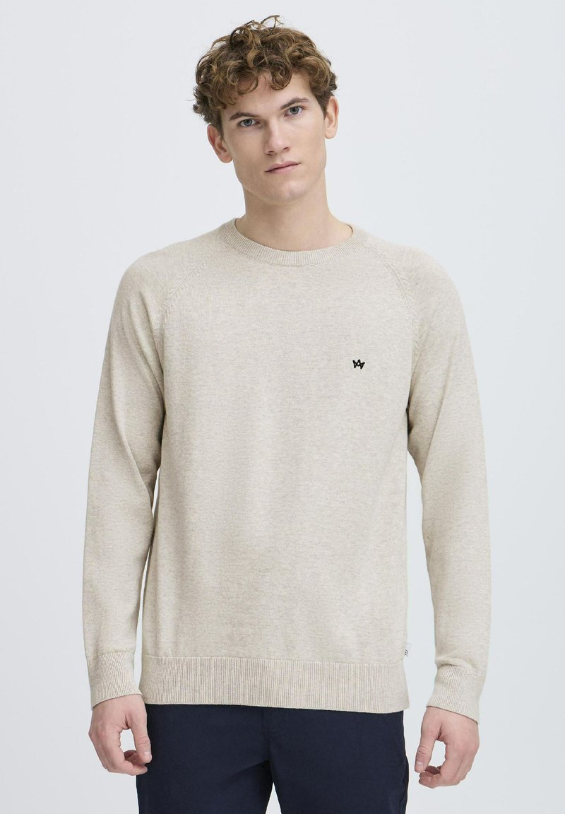Beige genser med rund hals, raglanerm og ribbestrikkede mansjetter. Har en liten svart logo på brystet, laget av myk teksturert stoff.