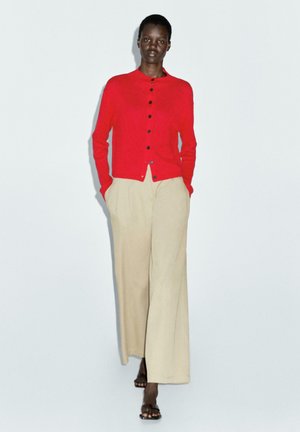 Cardigan rosso a maglia con scollatura tonda e bottoni neri, abbinato a pantaloni beige a gamba larga. La modella sta in piedi con le mani in tasca.