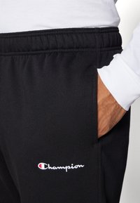 Melni sporta bikses ar elastīgu jostu, pievienoti sānu kabatas un balta "Champion" logo izšuvums uz kreisā augšstilba. Maiga auduma tekstūra.