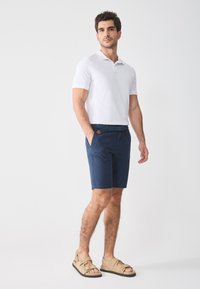 Navy blue shorts with a woven belt and tan accents, paired with a fitted white polo and light brown sandals. Smooth texture throughout.