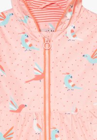 Světlé růžové deštivé sako s vzorem barevných ptáků a červenými puntíky. Má zipové zapínání a nařasený lem.