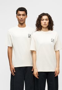 Βαμβακερές μπλούζες σε εκρού, με κοντά μανίκια και τετράγωνο γραφικό με το κείμενο «Flowers» σε μαύρο στο στήθος. Φοριούνται με χαλαρά σκουρόχρωμα παντελόνια.